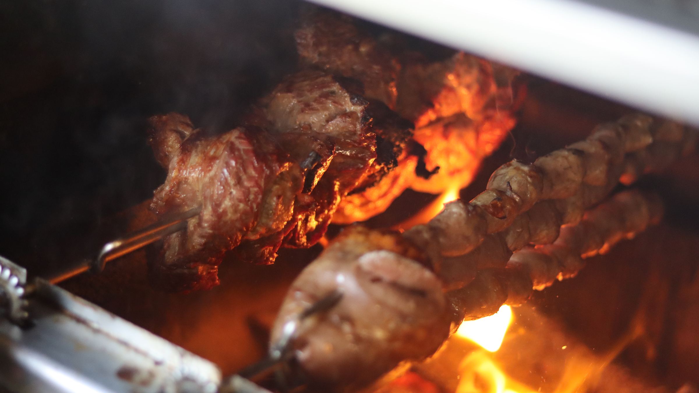 Espetos de carne assando sobre a brasa, com a chama subindo atrás