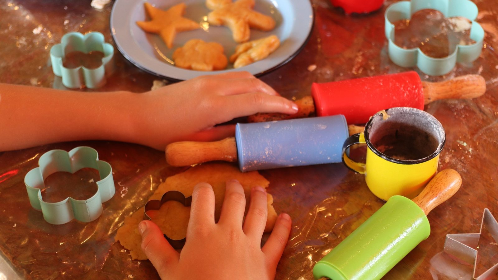 Mãos de criança cortando a massa com os cortadores de biscoito, entre rolos coloridos e as canecas de esmalte