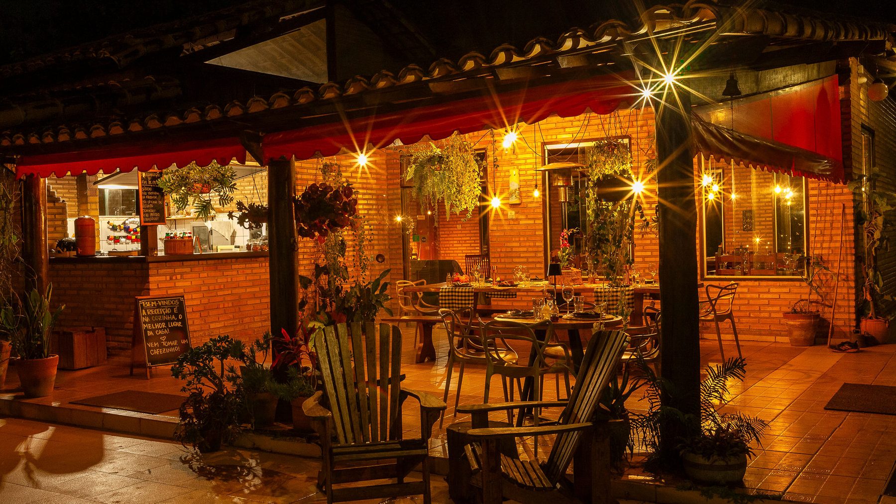 A varanda do restaurante acesa à noite, com as mesas postas, as luzes penduradas e as plantas
