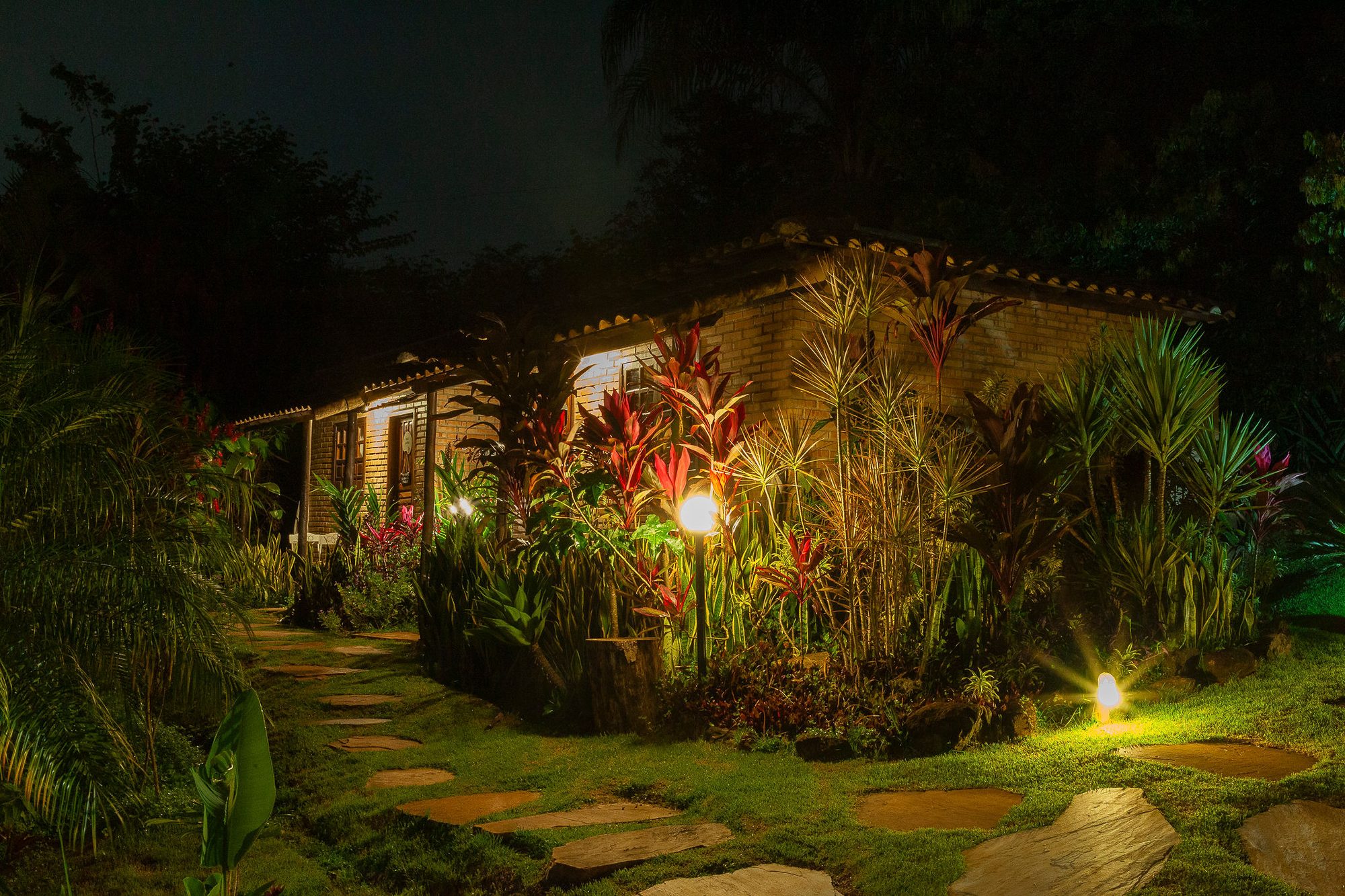 Os chalés acesos à noite, entre as folhagens vermelhas do jardim, com o caminho de pedras no gramado e as luminárias baixas
