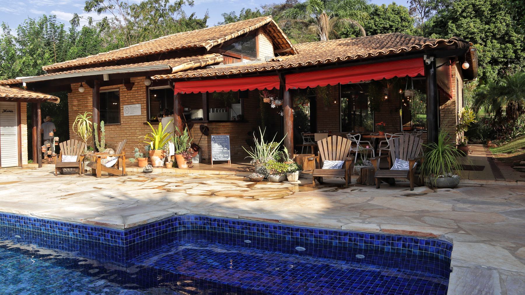A casa vista da beira da piscina, com o toldo vermelho sobre a varanda e as cadeiras de madeira no piso de pedra