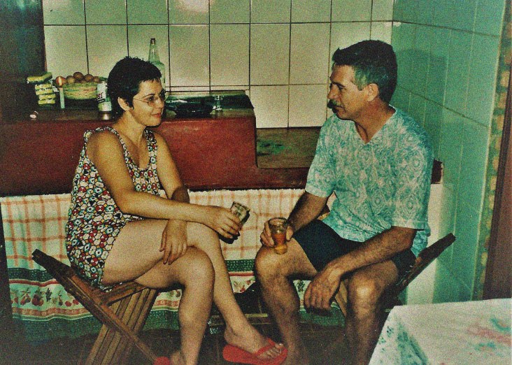 Luiz e Dagmar sentados frente a frente em cadeiras de madeira, na cozinha de azulejos, cada um com um copo na mão, conversando