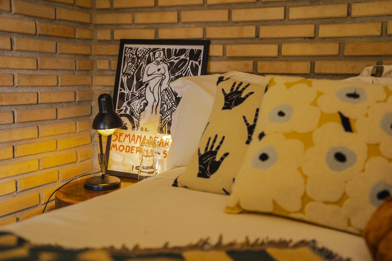 Cama do Chalé do Jacarandá contra a parede de tijolo, com almofadas estampadas e um quadro apoiado