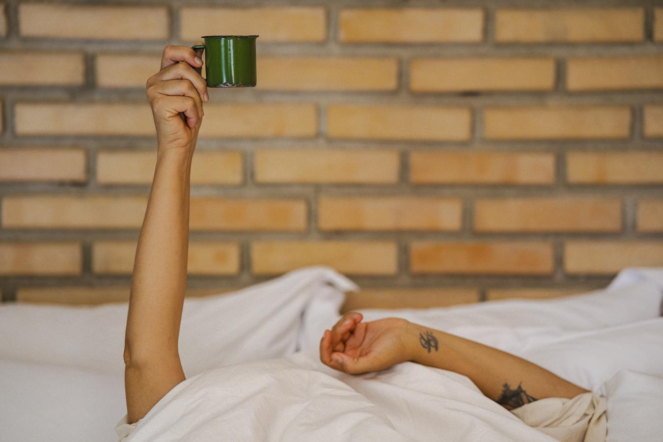 Uma mão erguendo a caneca verde acima do lençol branco, contra a parede de tijolo