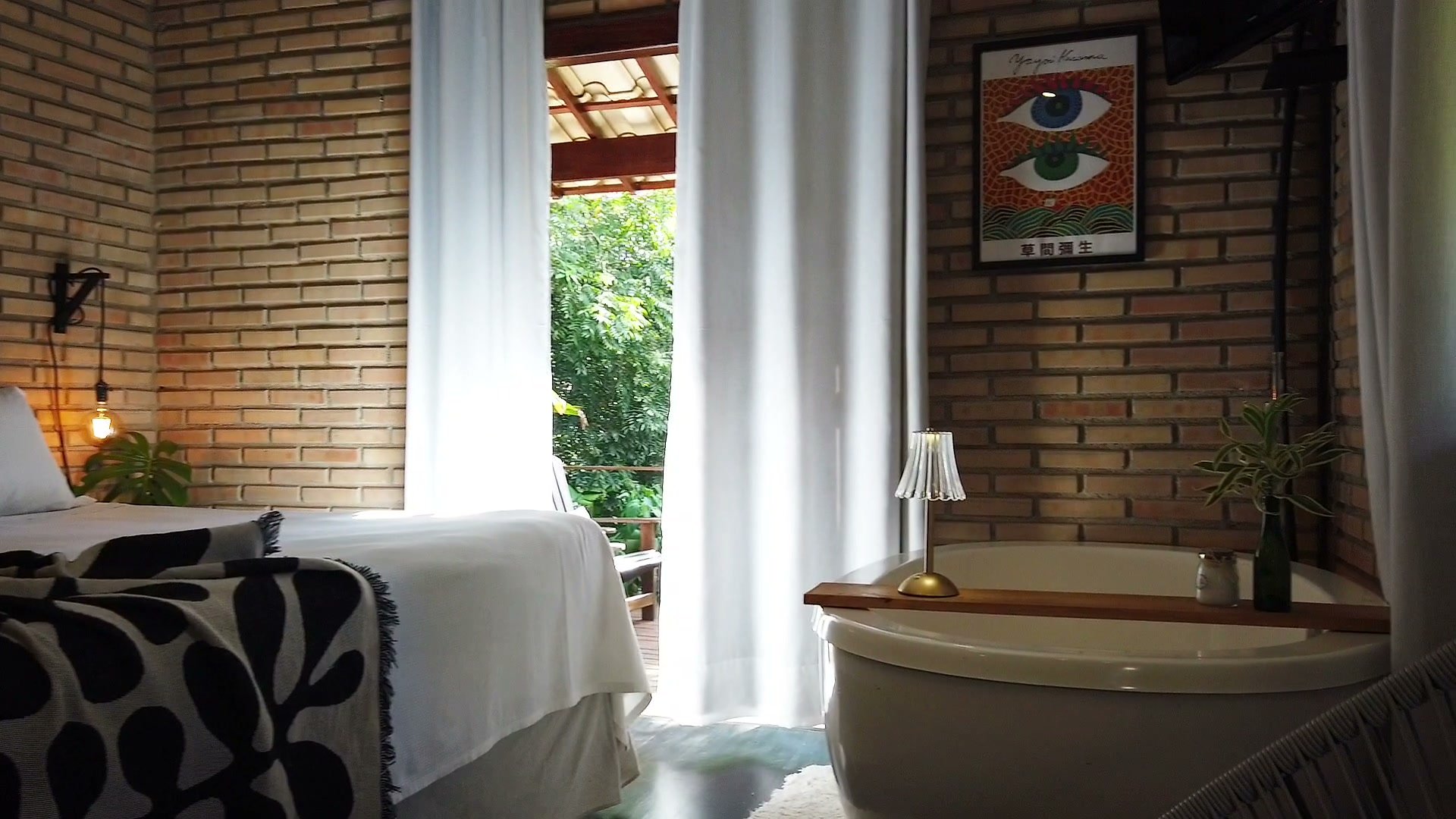 A banheira do Chalé Jacarandá Mata ao lado da cama, com as cortinas brancas abertas para o verde e a manta de folhas preta e branca
