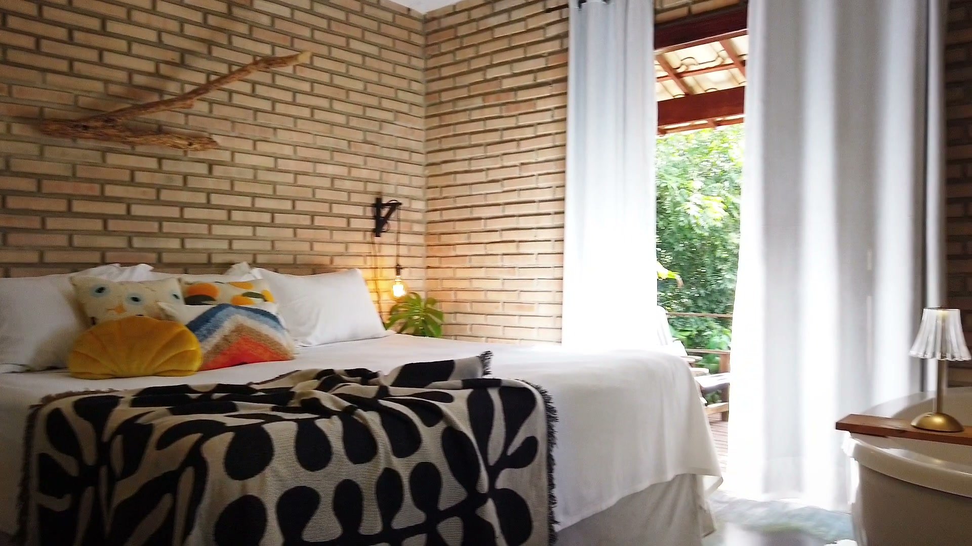 A cama com as almofadas coloridas e a manta preta e branca, ao lado da janela de cortinas brancas
