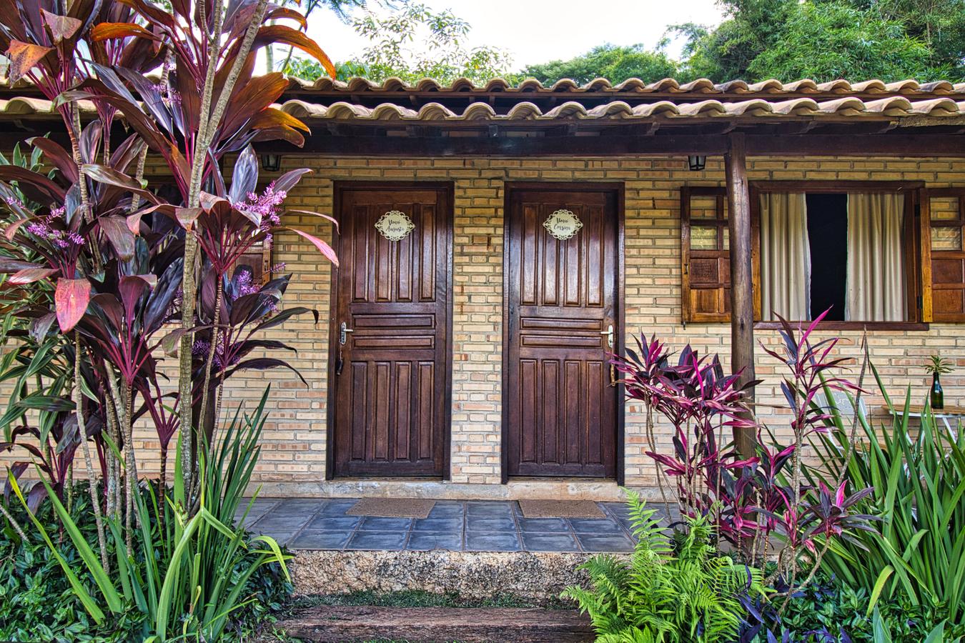 A fachada dos Chalés das Vovós: duas portas de madeira escura com as plaquinhas dos nomes, sob o telhado de telha, entre as folhagens