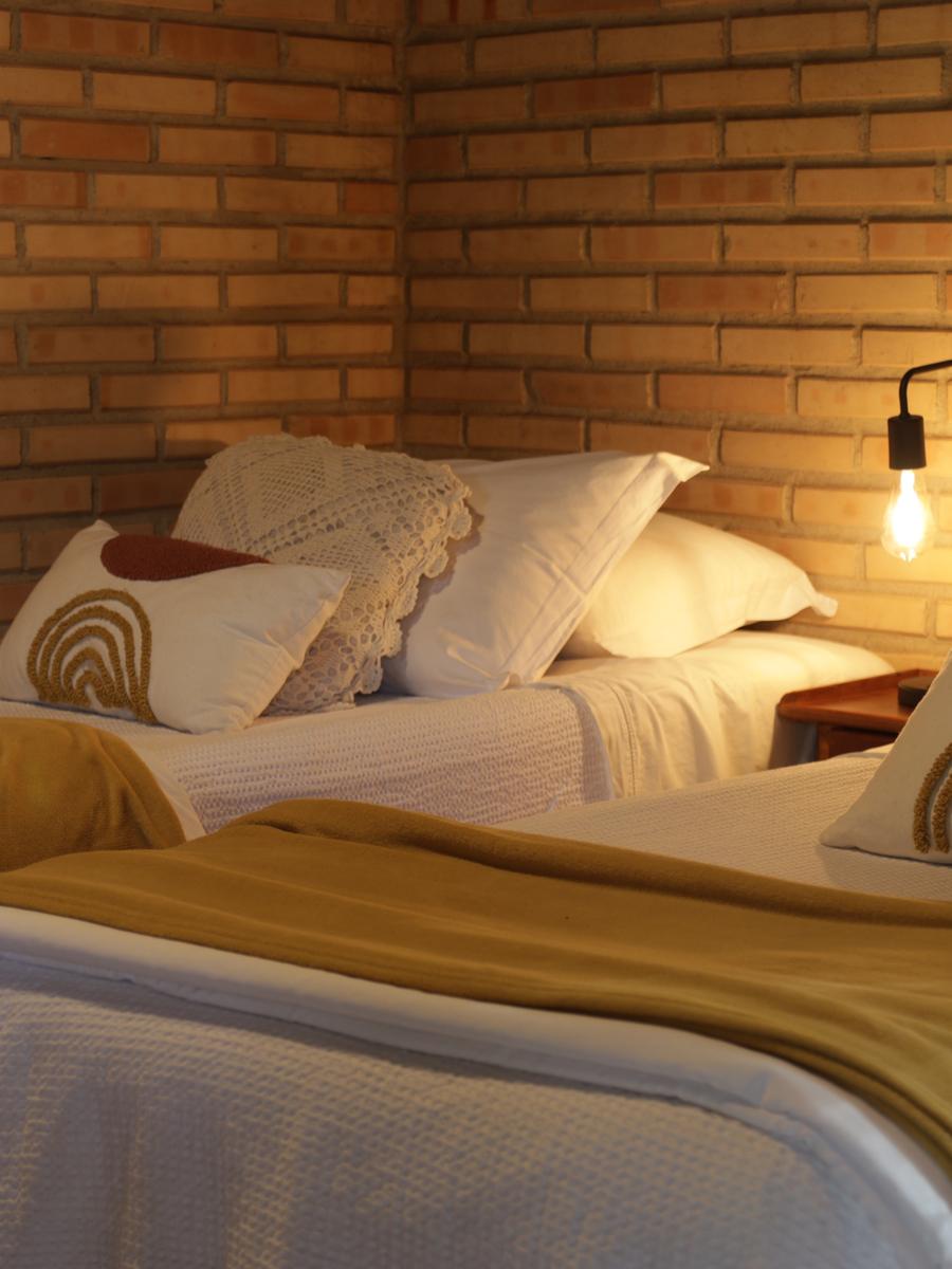 Quarto do Chalé das Vovós com cama de casal e cama auxiliar, parede de tijolo e luminária de leitura