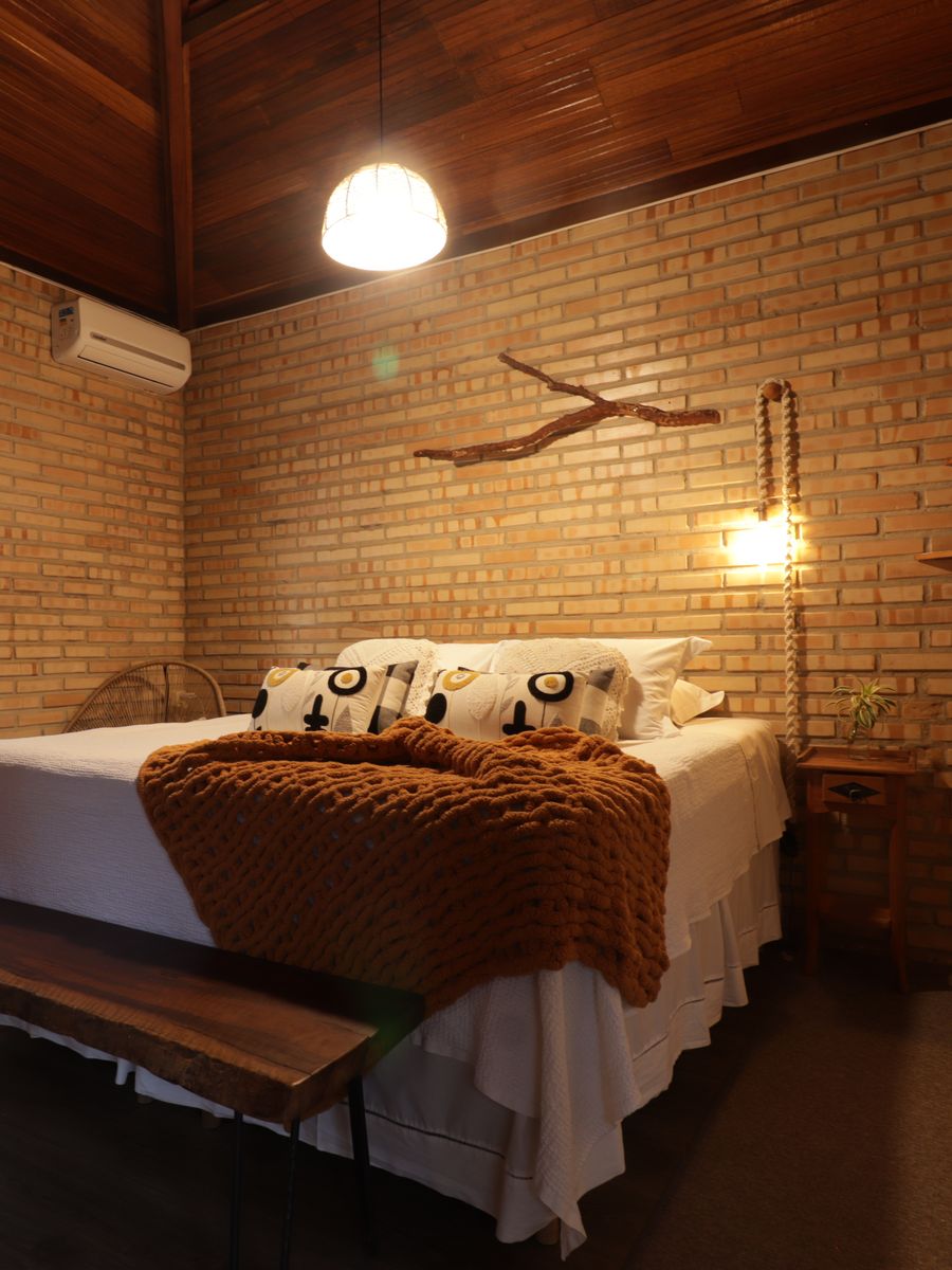 Quarto de casal com a manta de tricô cor de telha, o galho na parede de tijolo e a arandela acesa