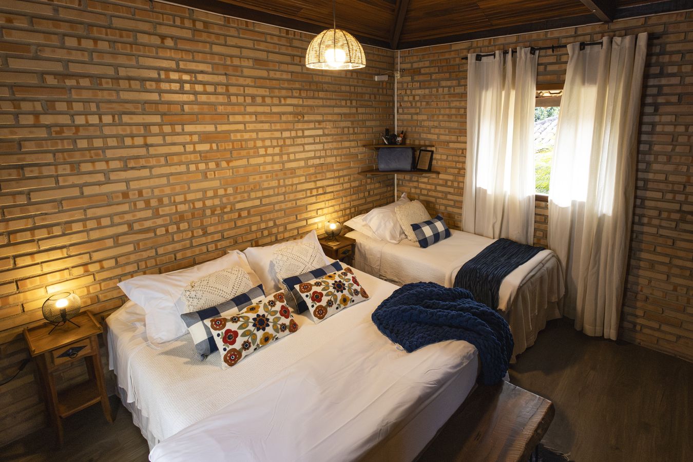 Quarto de duas camas com as almofadas bordadas de flores e a manta azul, a janela de cortina clara ao fundo