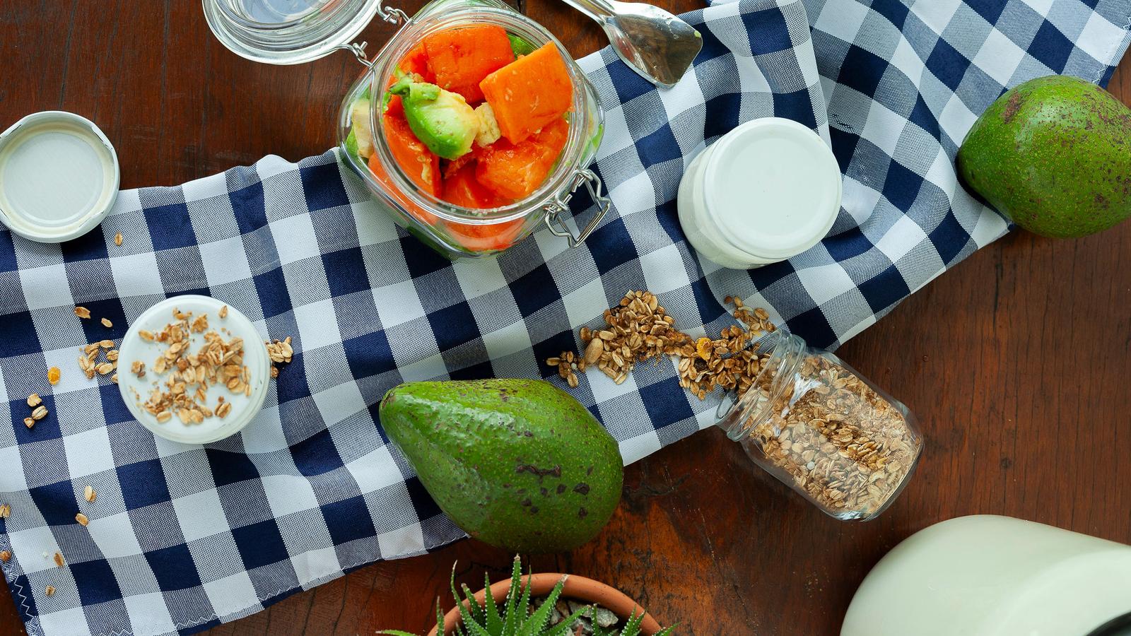 Mamão e abacate no vidro, iogurte, granola e os abacates inteiros sobre o pano xadrez, vistos de cima